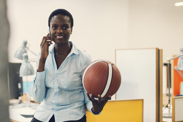Smiling African businesswoman sitting on desk with basketball ball and talking on the phone in modern office. Front view, selective focus to her smiling face.